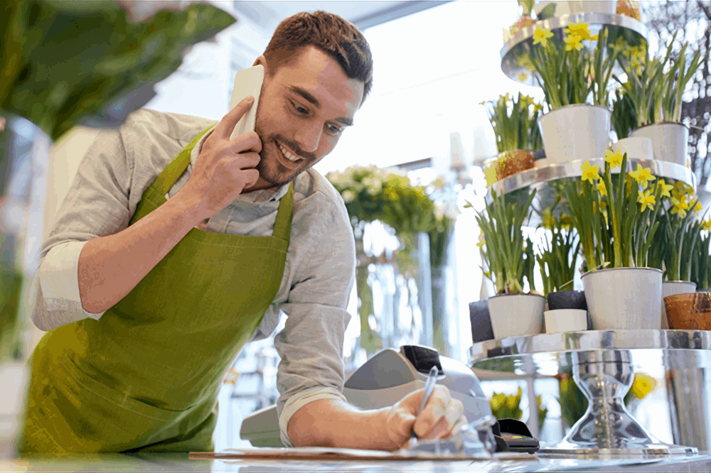 multiple pots of flowers sit in the background as a happy florist talks on a cellphone while taking notes on a counter