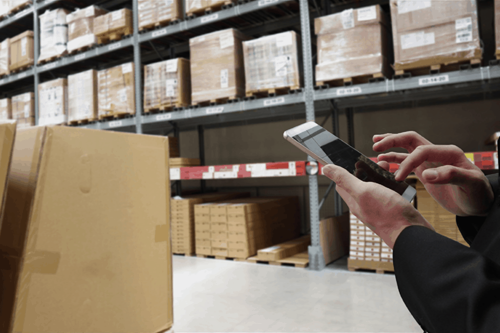 a warehouse employee types on a mobile phone in front of shelves filled with boxes